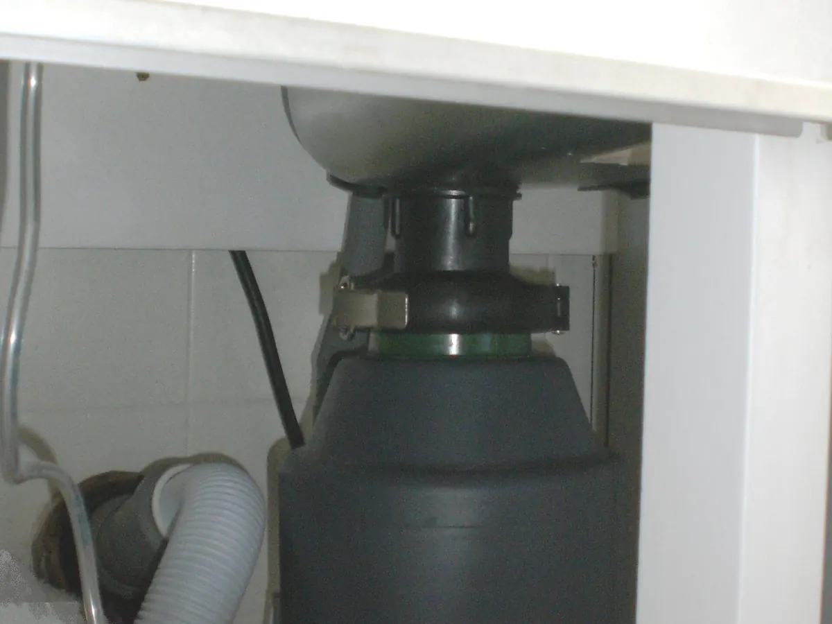 A garbage disposer mounted under a white kitchen sink, with its drain hose on the left